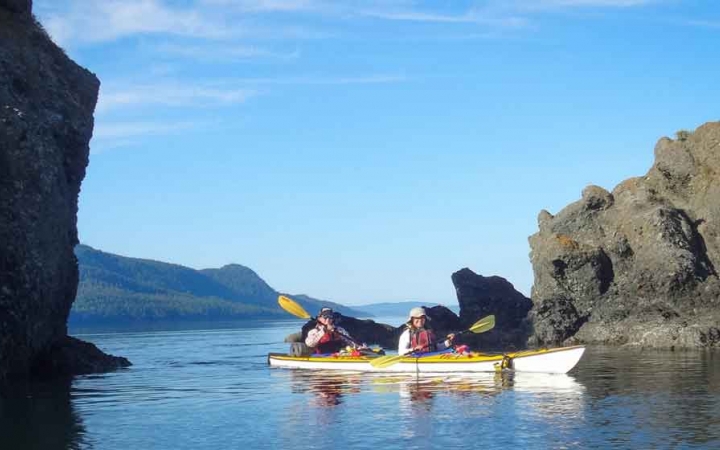 two people paddle a kayak in calm blue water between two rock formations in the pacific northwest